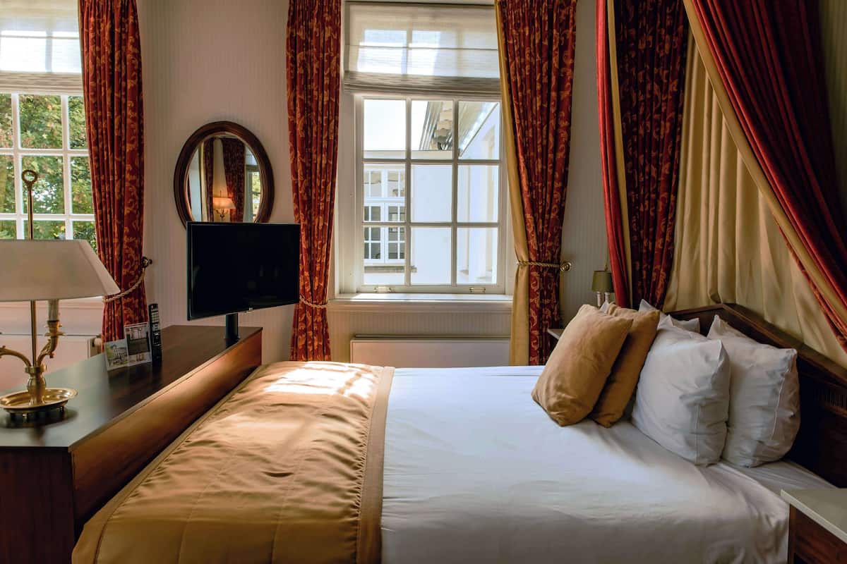 A hotel room with a bed, a desk with a lamp, a television, and a window with curtains.
