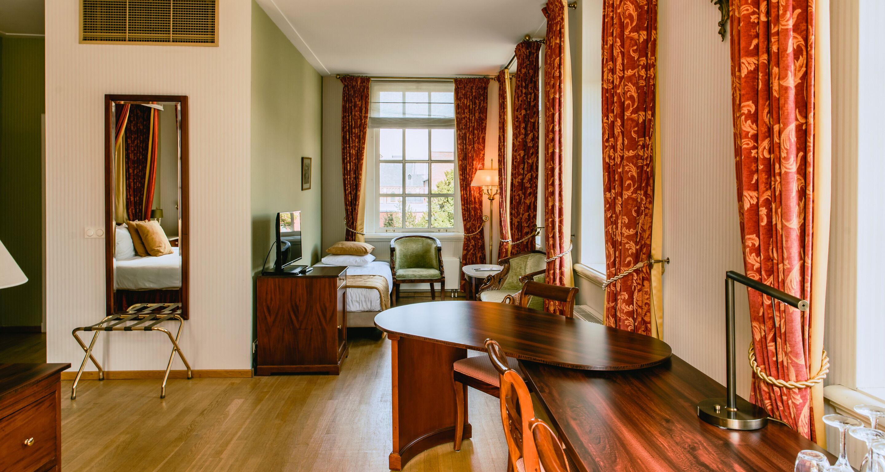 A hotel room with a wooden dining table, two chairs, a small side table, a green armchair, a wooden cabinet, and a bed with a headboard.