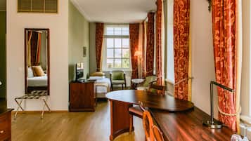 A hotel room with a wooden dining table, two chairs, a small side table, a green armchair, a wooden cabinet, and a bed with a headboard.