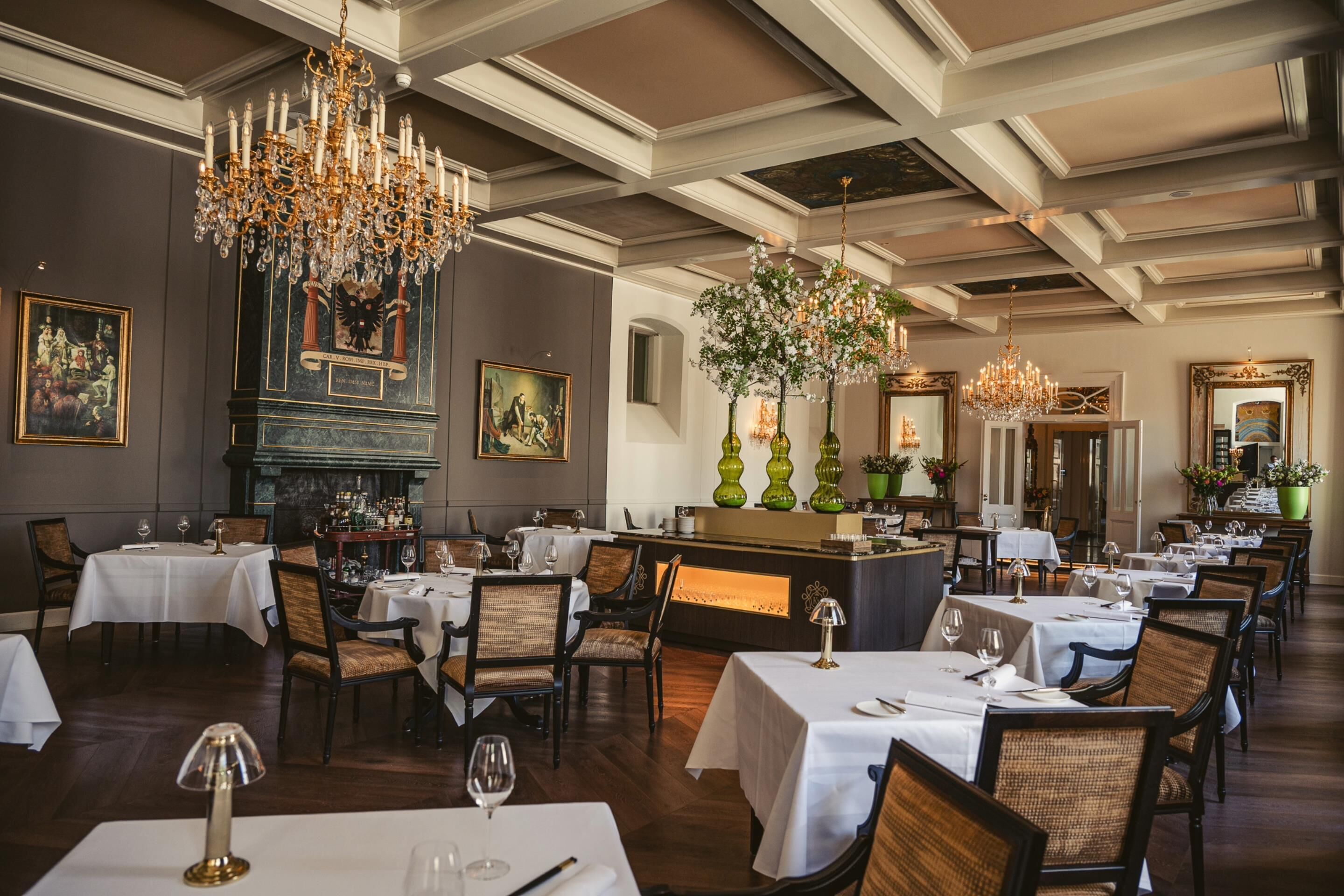 Uma sala de jantar formal, com lustres, mesas postas e um espaço de bar.