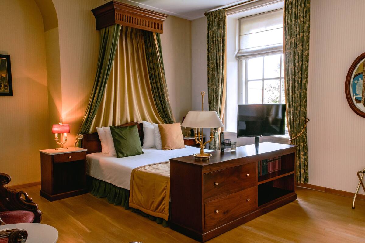 A bedroom with a bed, a nightstand, a dresser, a television, and a window with curtains.