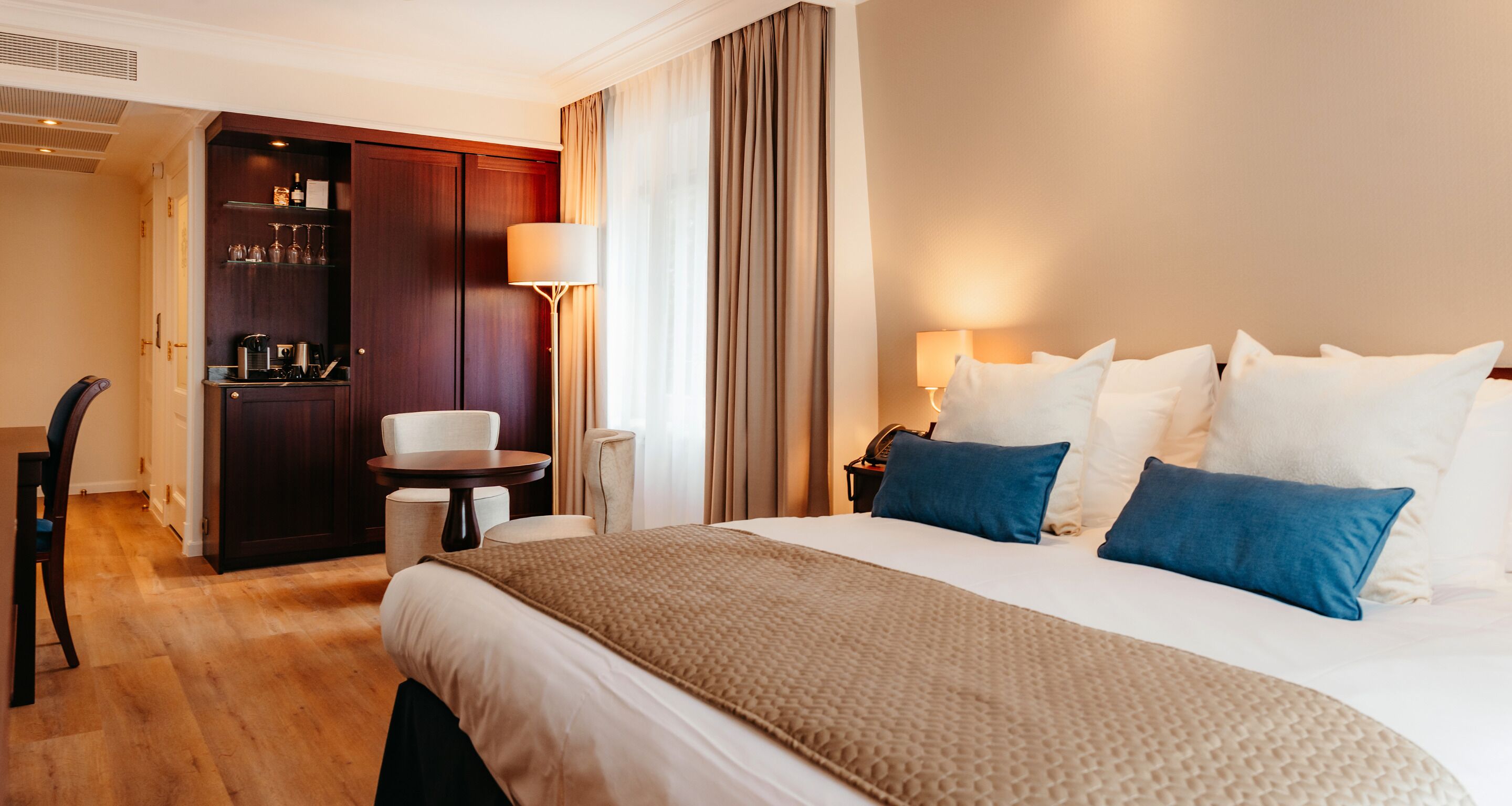 A hotel room with a large bed, a desk, a chair, a minibar, and a lamp.
