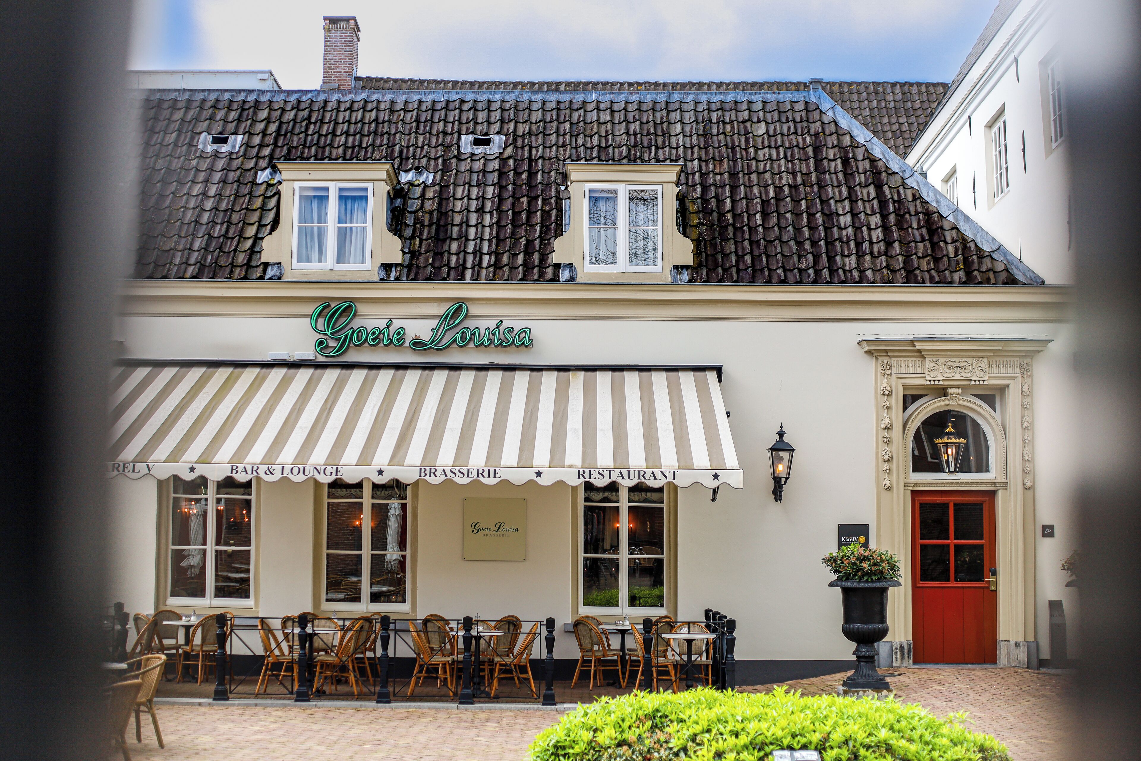 Een gebouw met een bord waarop 'Goie Louisa' staat, en een gestreepte luifel. Er staan buiten tafels en stoelen, en een rode deur.