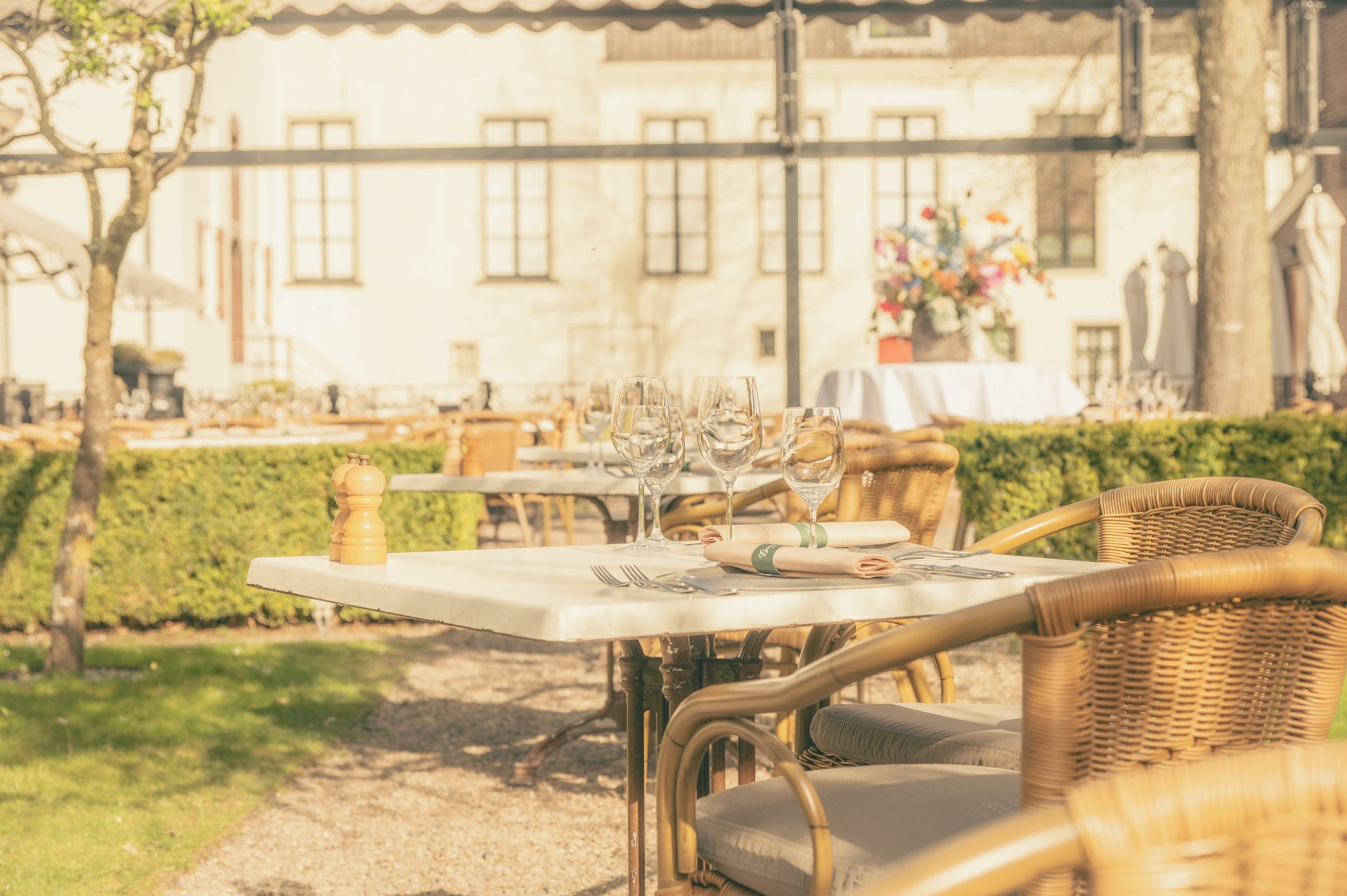 An outdoor dining area with tables set for two, featuring wicker chairs and a white tablecloth.