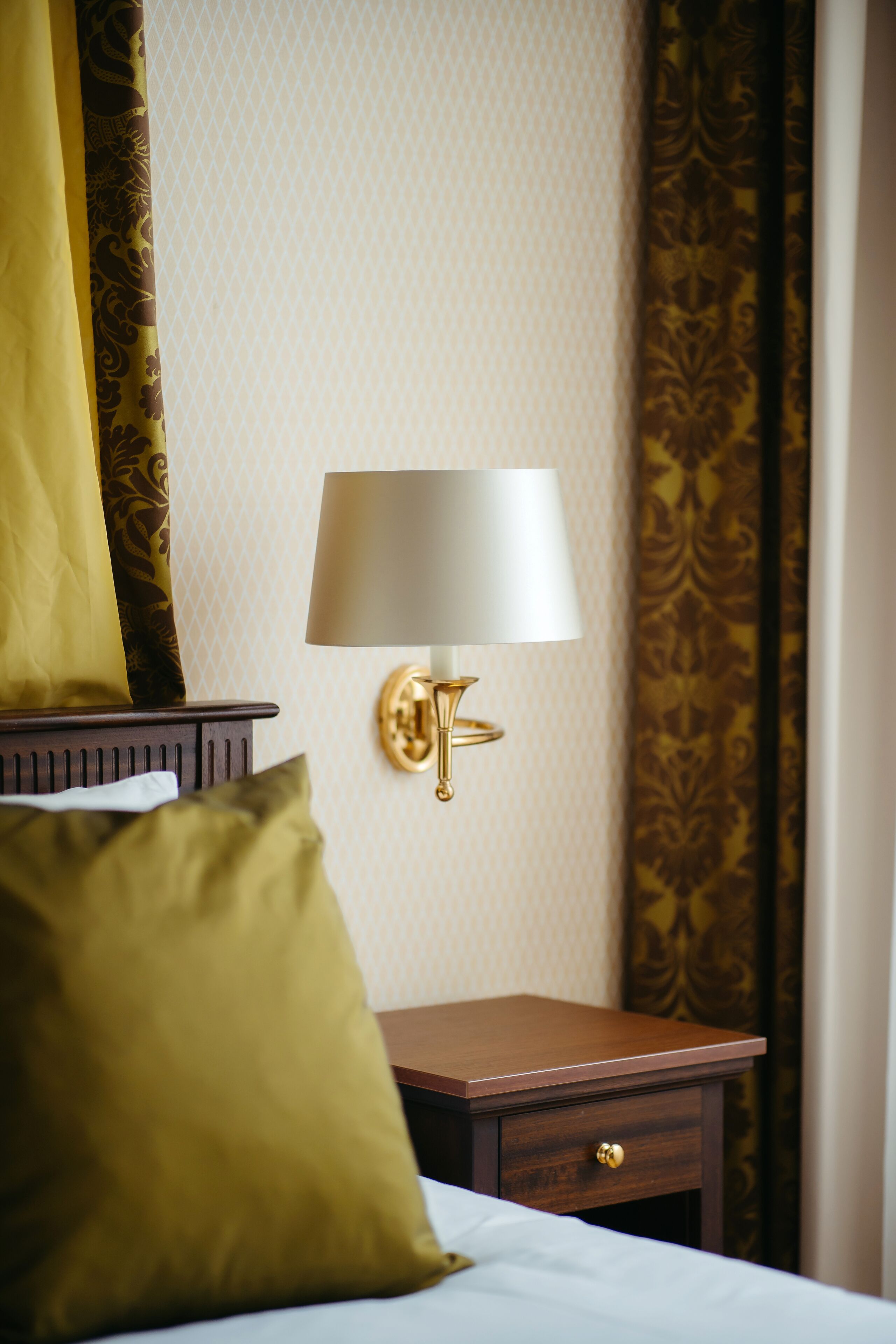 A neatly made bed with a green pillow, a bedside table with a lamp, and patterned curtains.