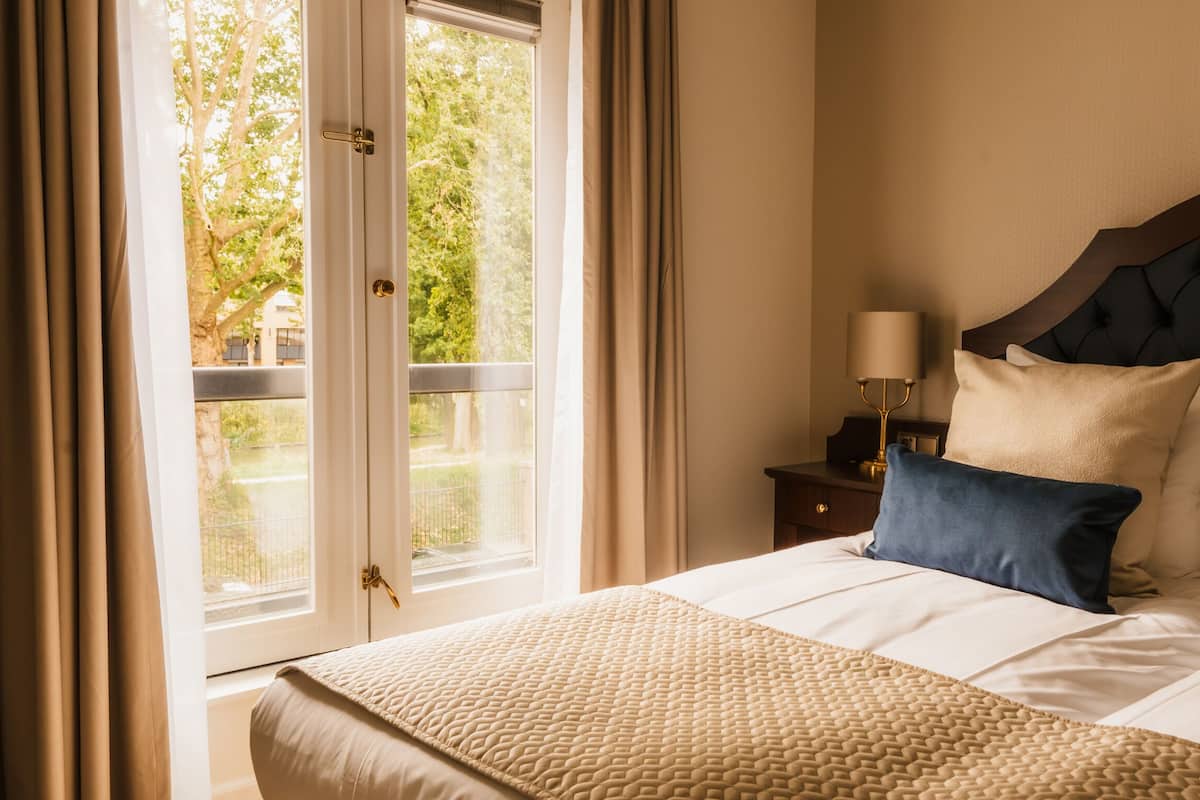 A hotel room with a large bed, a nightstand with a lamp, and a view of greenery through a window.