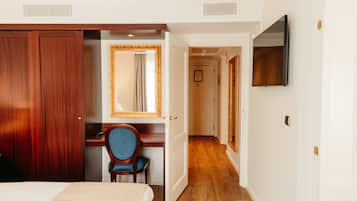 A hotel room with a wooden wardrobe, a desk with a chair, a large mirror, and a television on the wall.