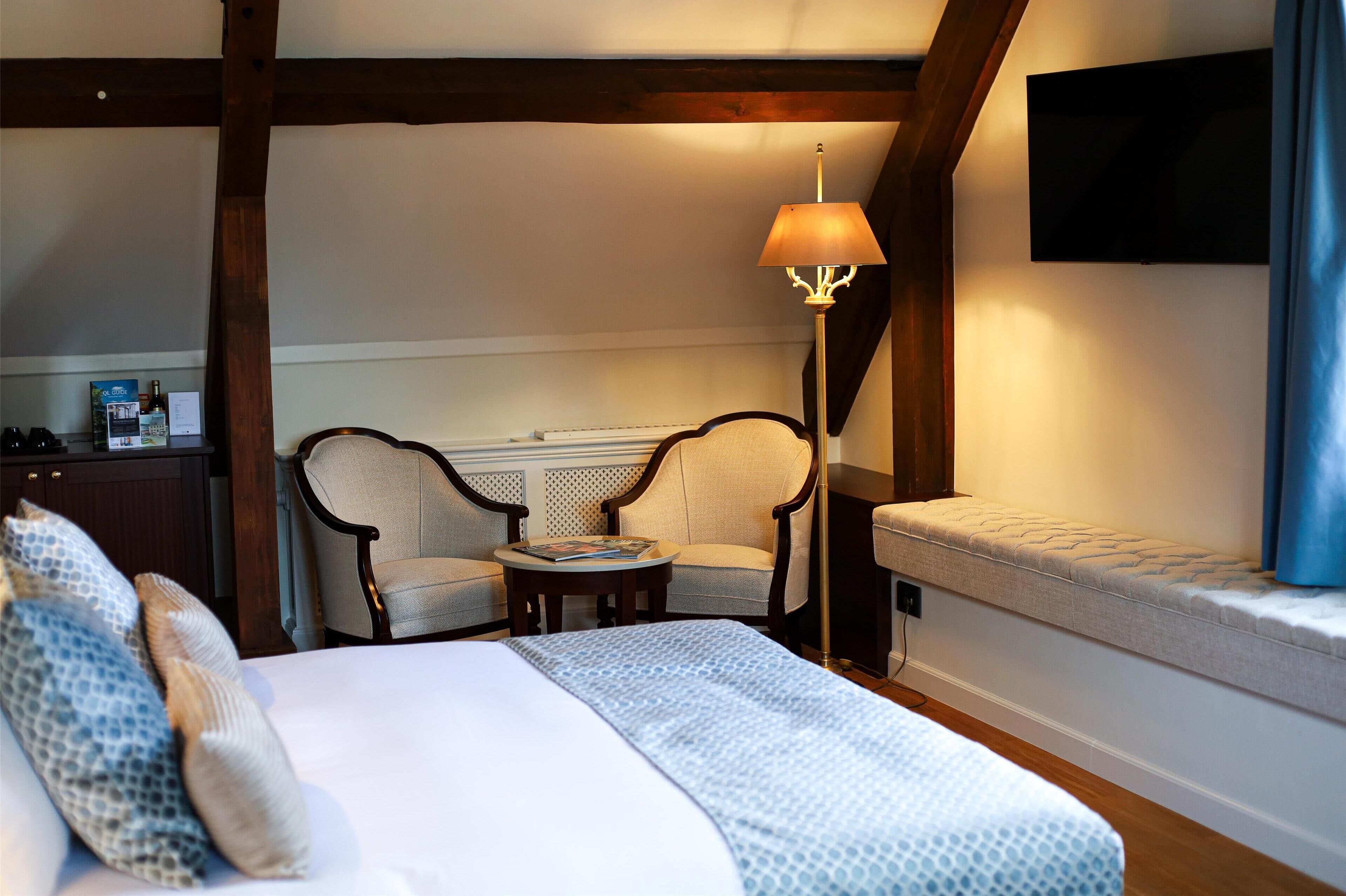 Habitación de hotel con una cama, dos sillones, una mesita, una lámpara, un televisor y un banco.
