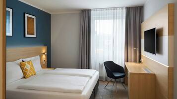 A hotel room with a bed, a desk, a chair, a TV, and two framed pictures on the wall.
