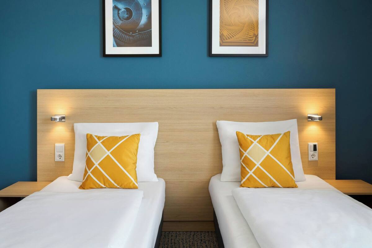 A hotel room with two single beds, each with a white mattress and a yellow pillow with a white pattern. The headboard is wooden, and there are wall-mounted light fixtures on either side.