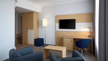 Una habitación de hotel moderna con un escritorio de madera, dos sillas azules, un televisor de pantalla plana en la pared y una pequeña kitchenette.