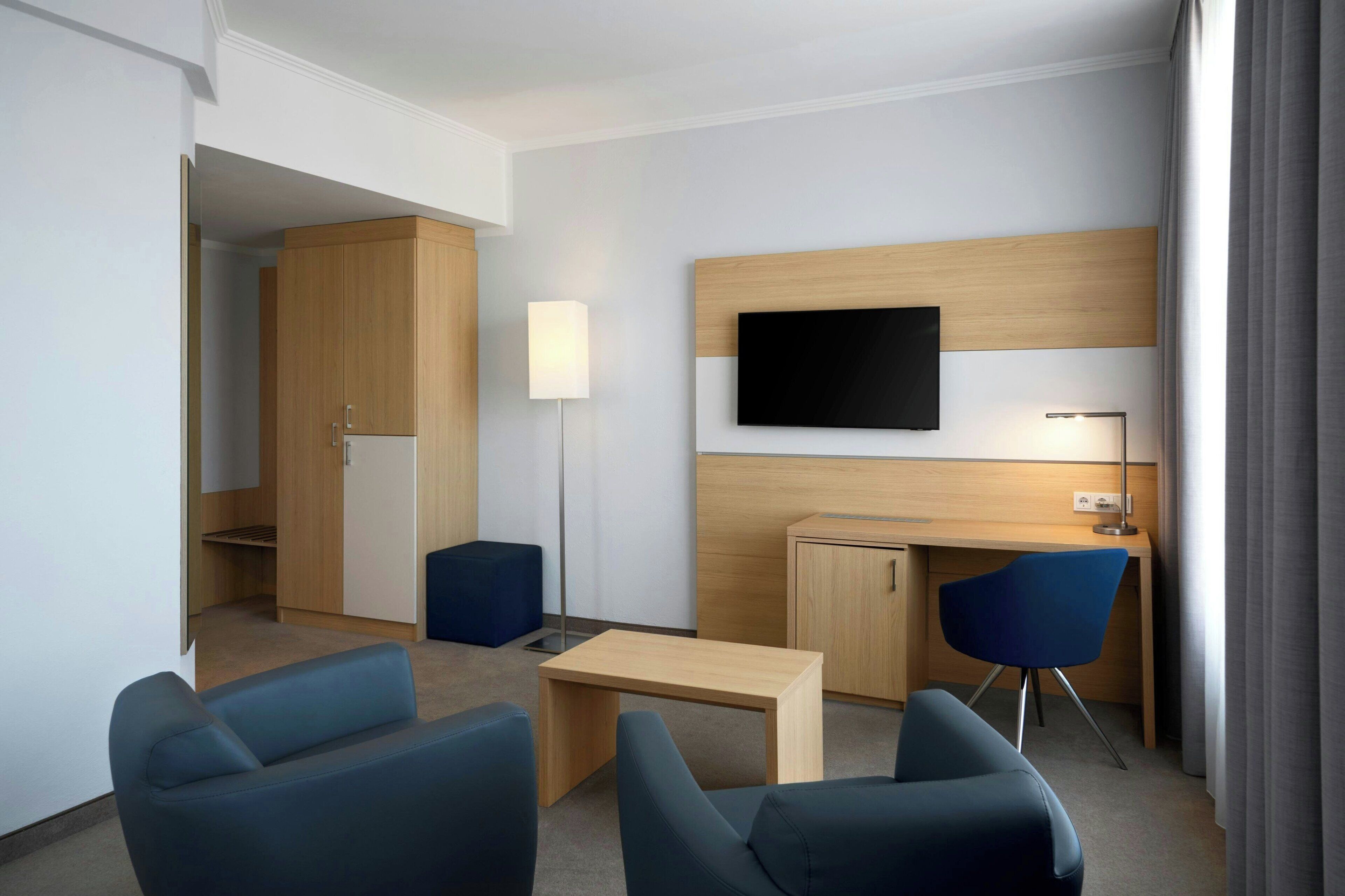 Een moderne hotelkamer met een houten bureau, twee blauwe stoelen, een flatscreen televisie aan de muur, een bedlamp en een kledingkast.
