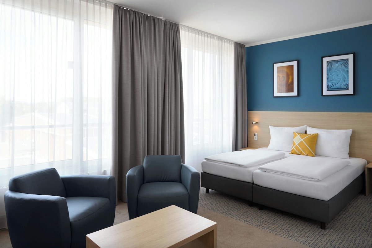 A hotel room with a bed, two chairs, a small table, and a large window with curtains.