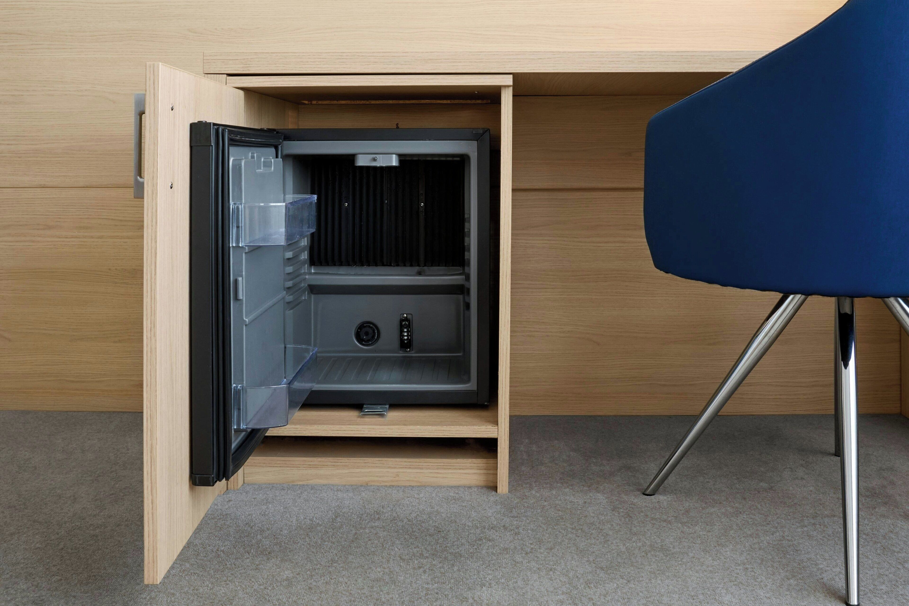 A compact, built-in safe with a transparent door, located under a wooden desk with a blue chair.