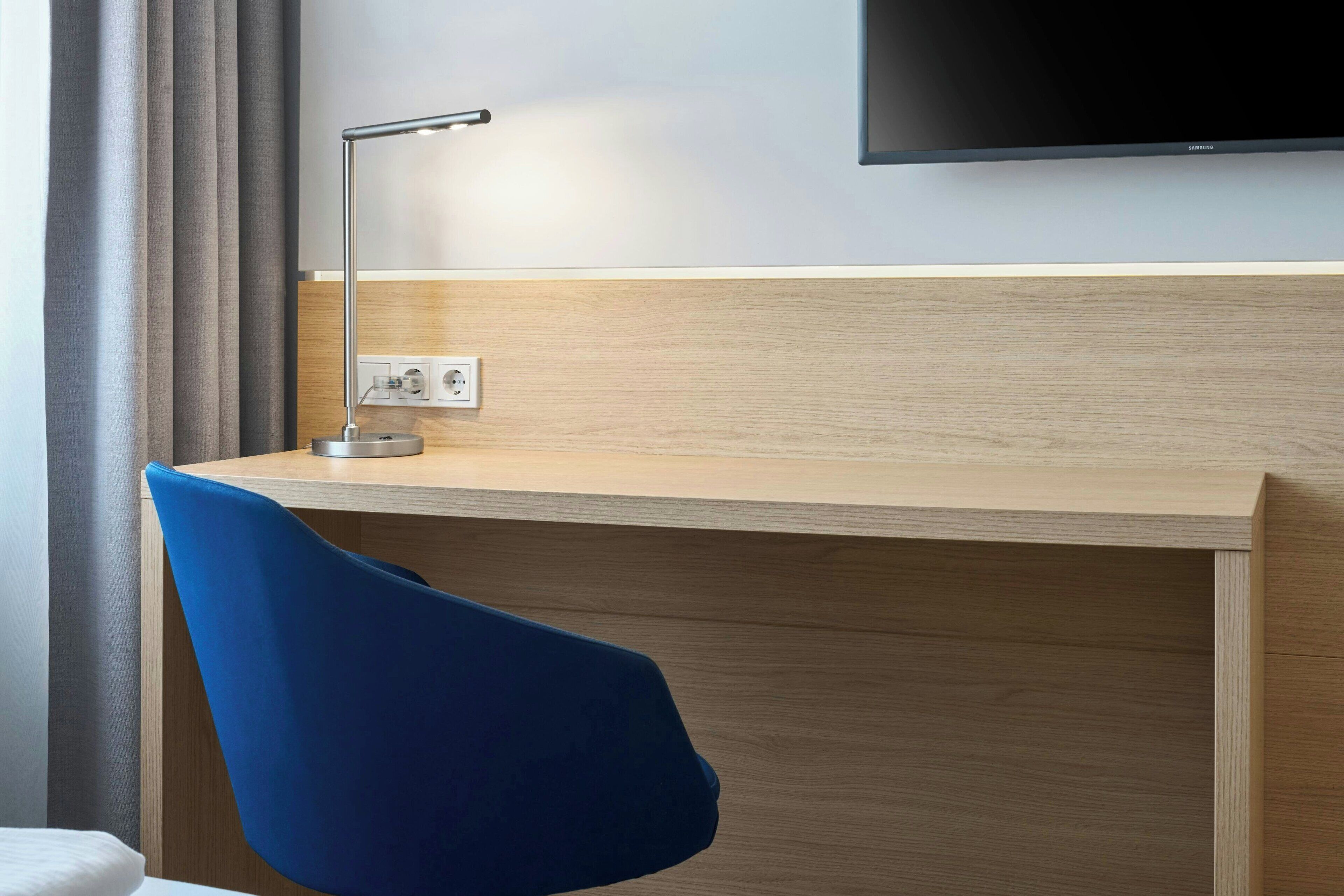 Habitación de hotel con escritorio de madera, una silla azul, un televisor de pantalla plana montado en la pared y un enchufe en el escritorio.