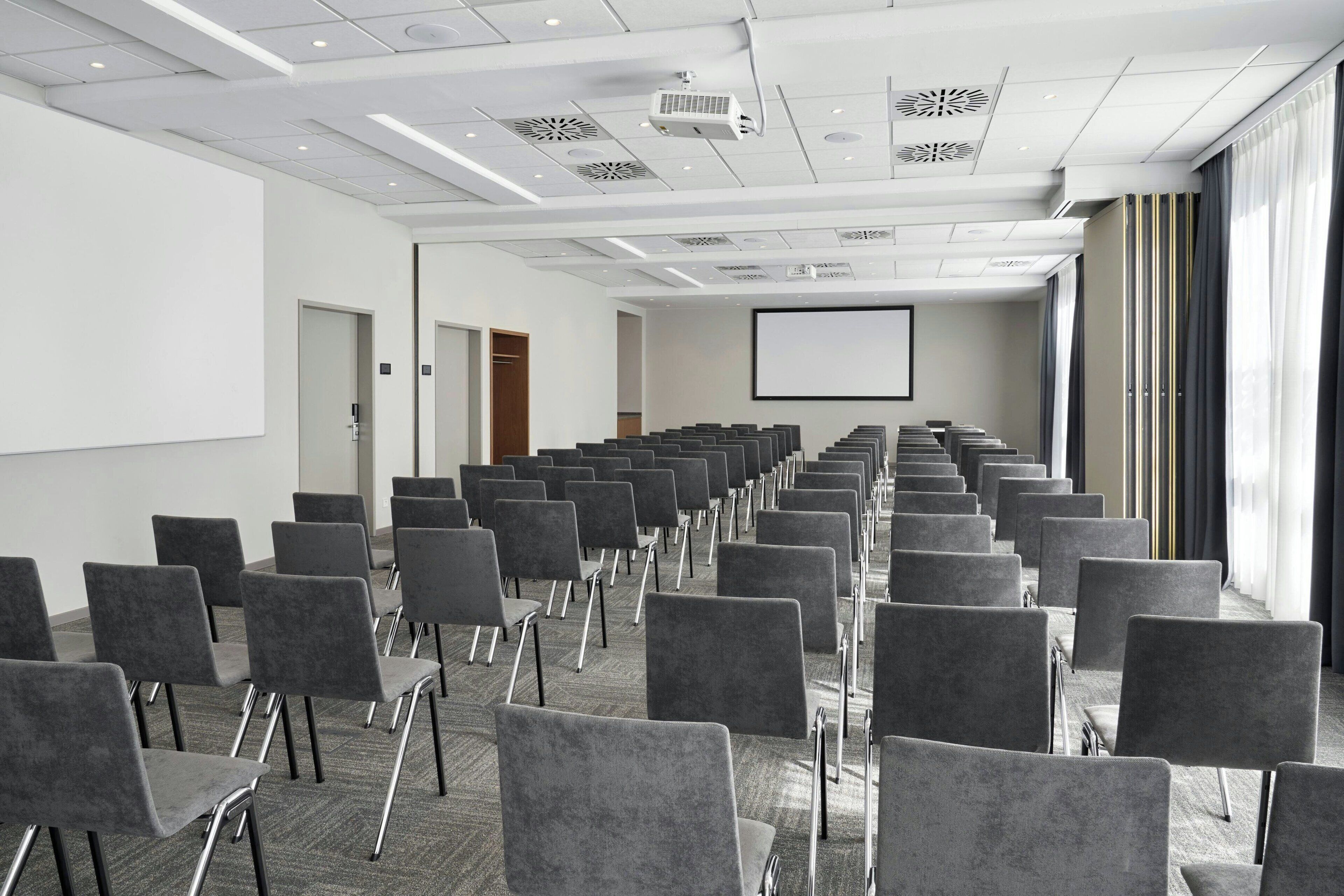 Un salón de conferencias con filas de sillas grises, una pantalla de proyector y un amplio ventanal con cortinas.