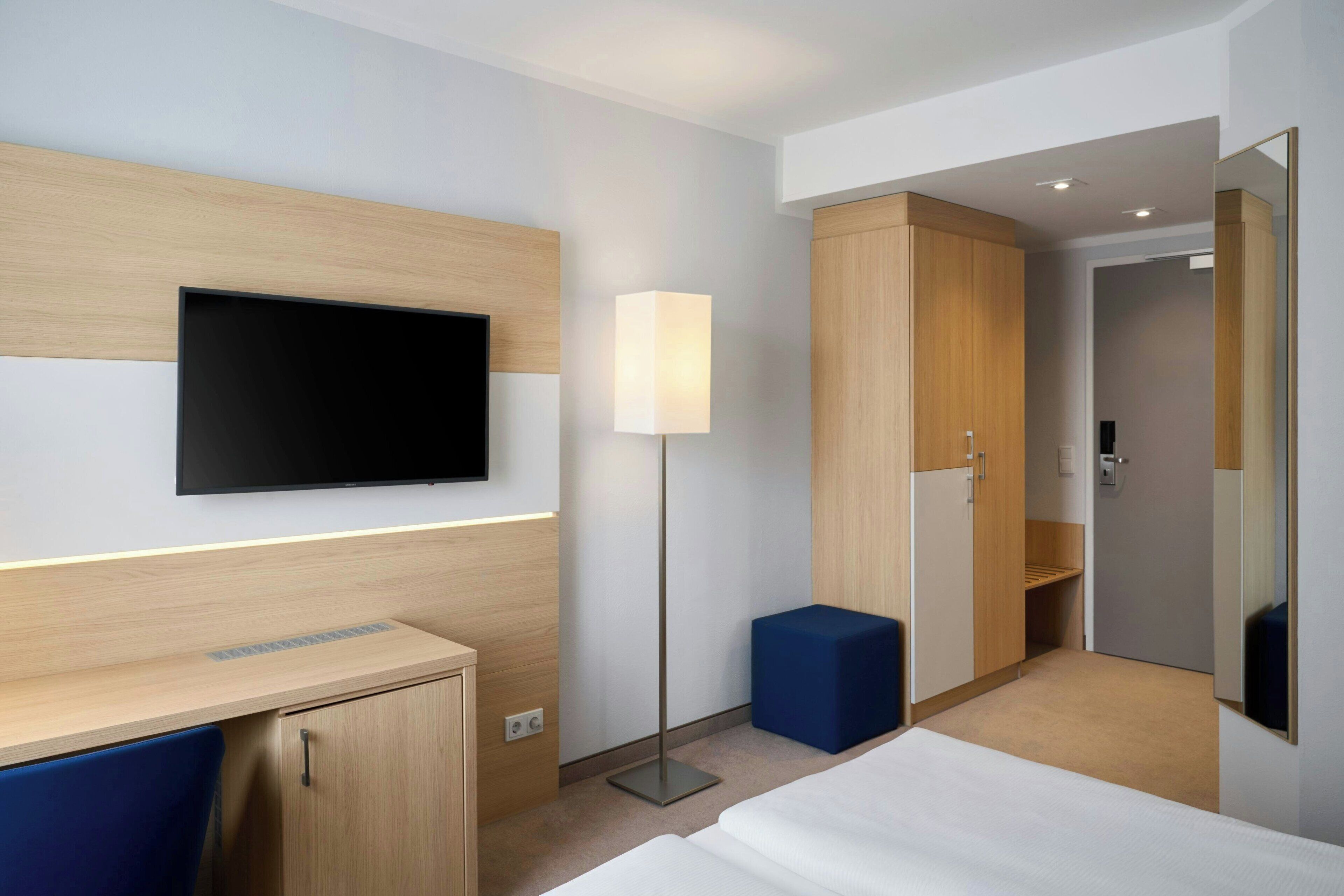 Una habitación de hotel con cama, escritorio, televisor montado en la pared, armario y lámpara de noche.