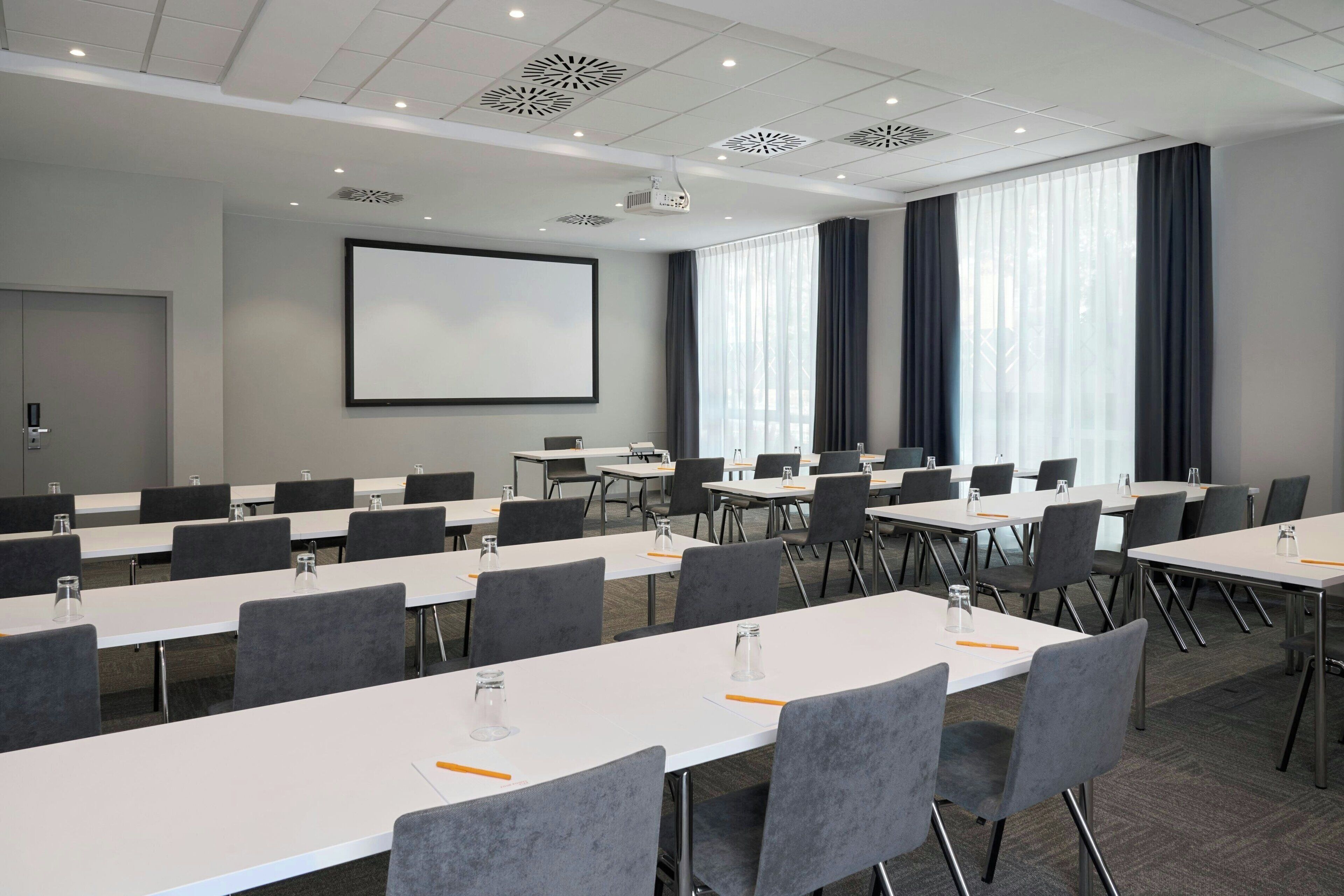 Une salle de conférence avec des tables blanches, des chaises grises, un écran de projection et de grandes fenêtres équipées de rideaux.