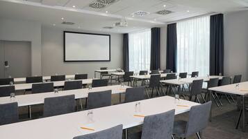Une salle de conférence avec des tables blanches, des chaises grises, un écran de projection et de grandes fenêtres équipées de rideaux.