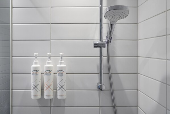 A bathroom with white tiled walls, a showerhead, and three bottles of ACTIV body wash and conditioner. - Four Points Flex by Sheraton Goettingen (Goettingen)