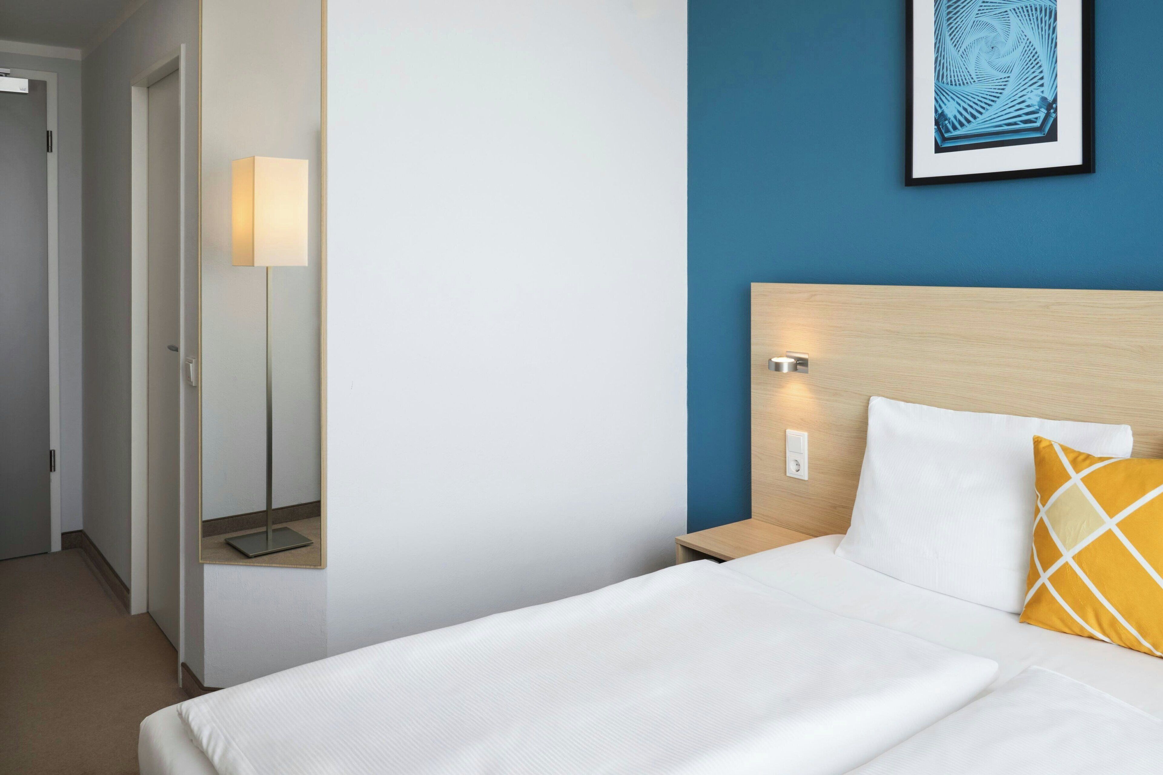 A hotel room with a bed, a nightstand, a bedside lamp, a wall-mounted artwork, and a door to another room.