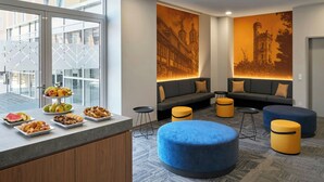 Un área de lounge moderna con una disposición de asientos, una presentación de frutas y un mural con vista a la ciudad en la pared.