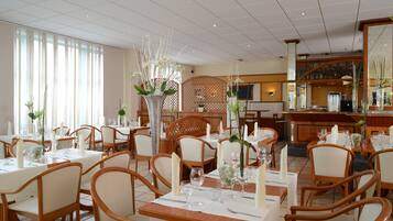 Un restaurante con mesas y sillas de madera, manteles blancos y centros de mesa florales.