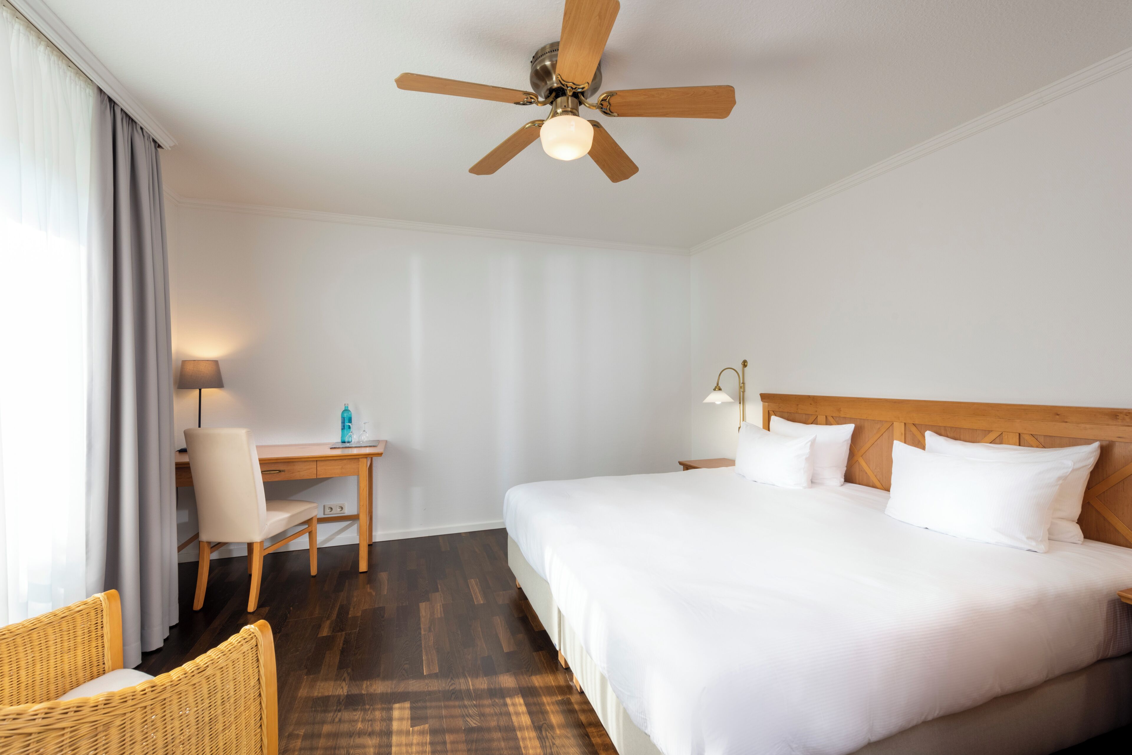 A hotel room with a bed, a desk, a chair, a ceiling fan, and a lamp.