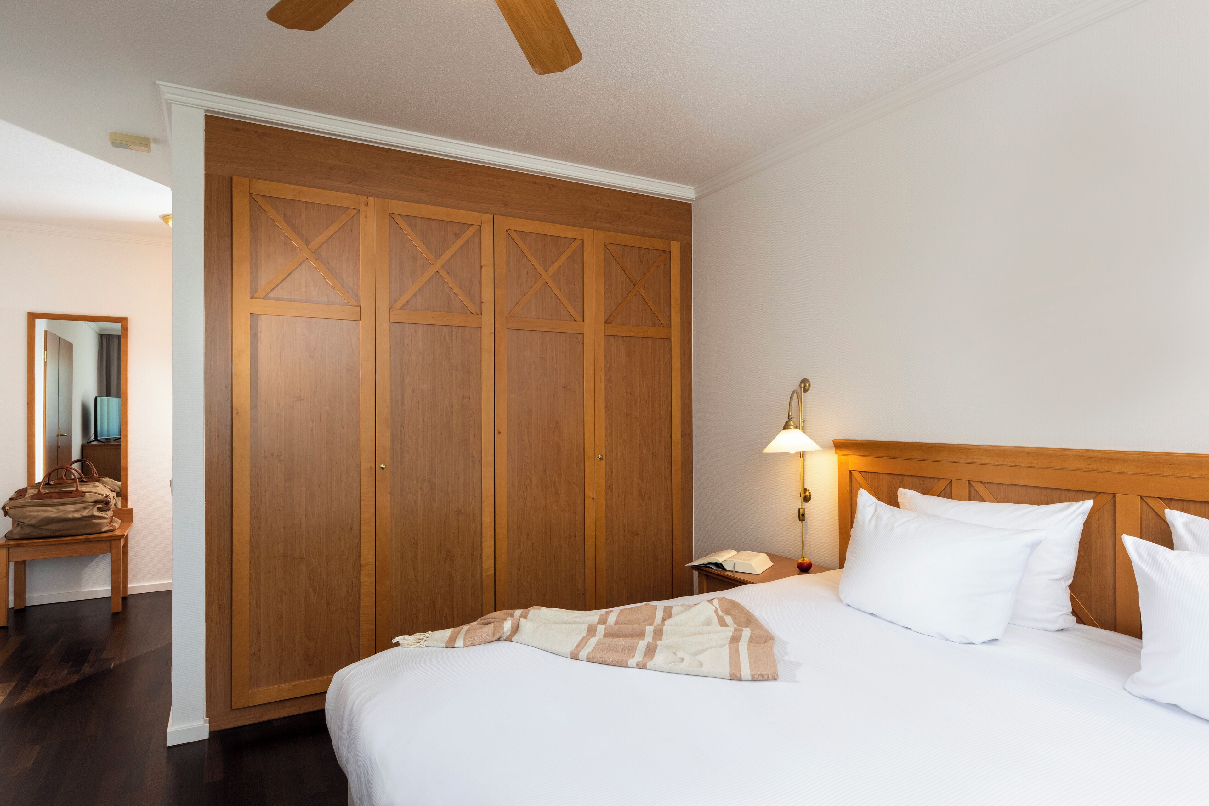 Habitación de hotel con un armario grande de madera, una cama con ropa de cama blanca, una lámpara de noche y un cabecero de madera.