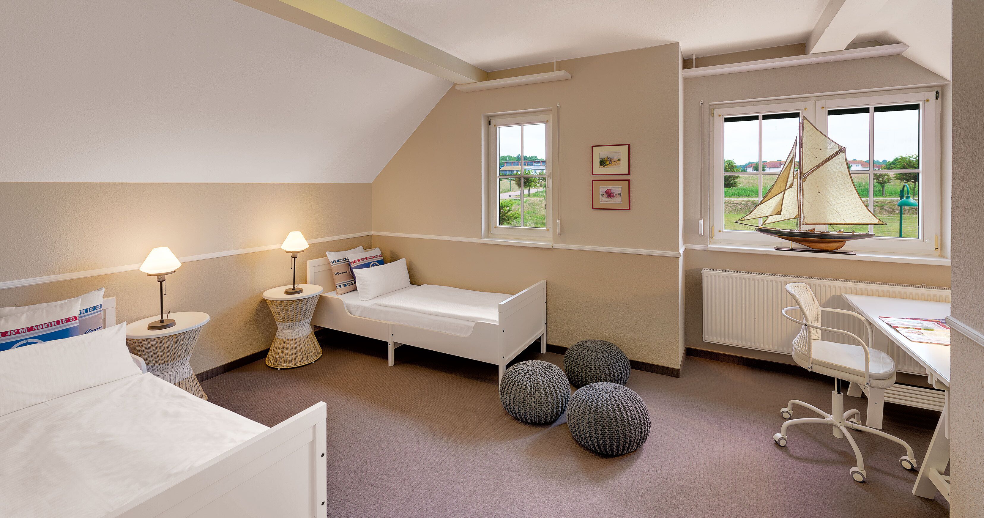 A bedroom with a bed, bedside lamp, pillows, a small round table, a chair, a radiator, and a window with a sailboat view.
