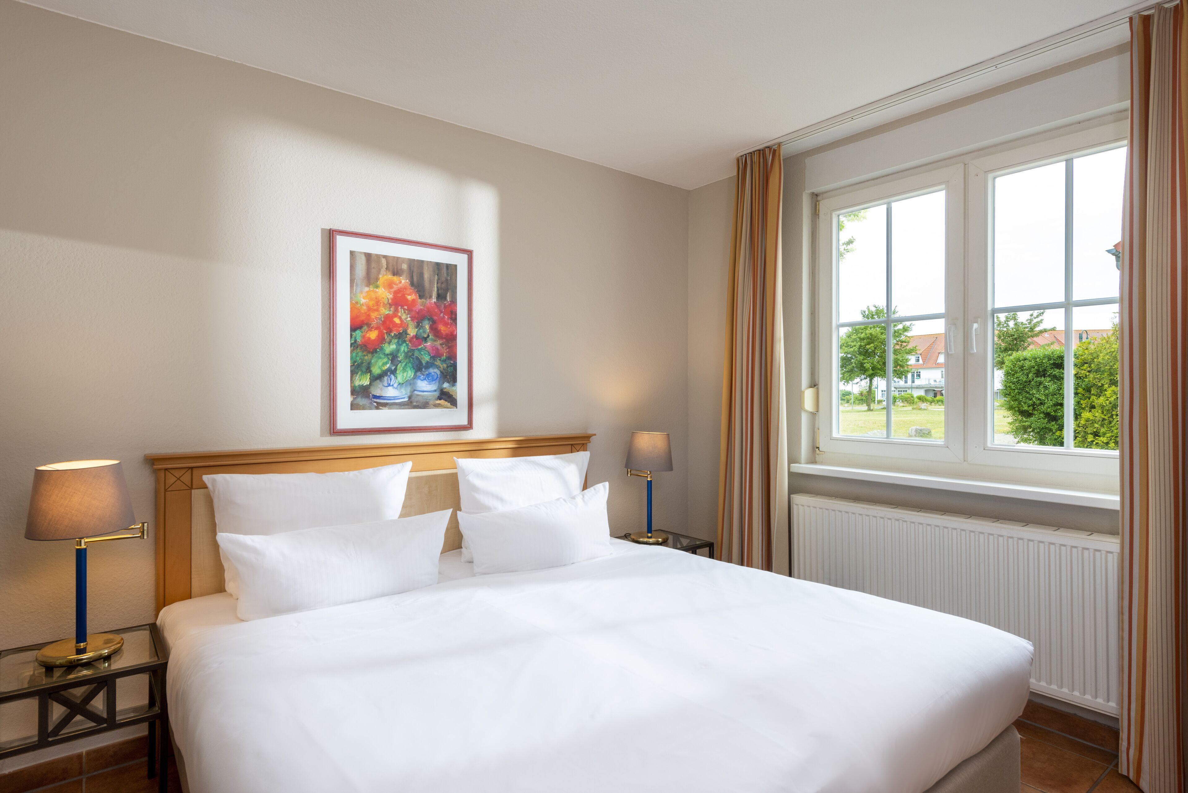 Una habitación de hotel con una cama grande, dos mesitas de noche con lámparas, un cuadro floral enmarcado en la pared y una ventana con cortinas.