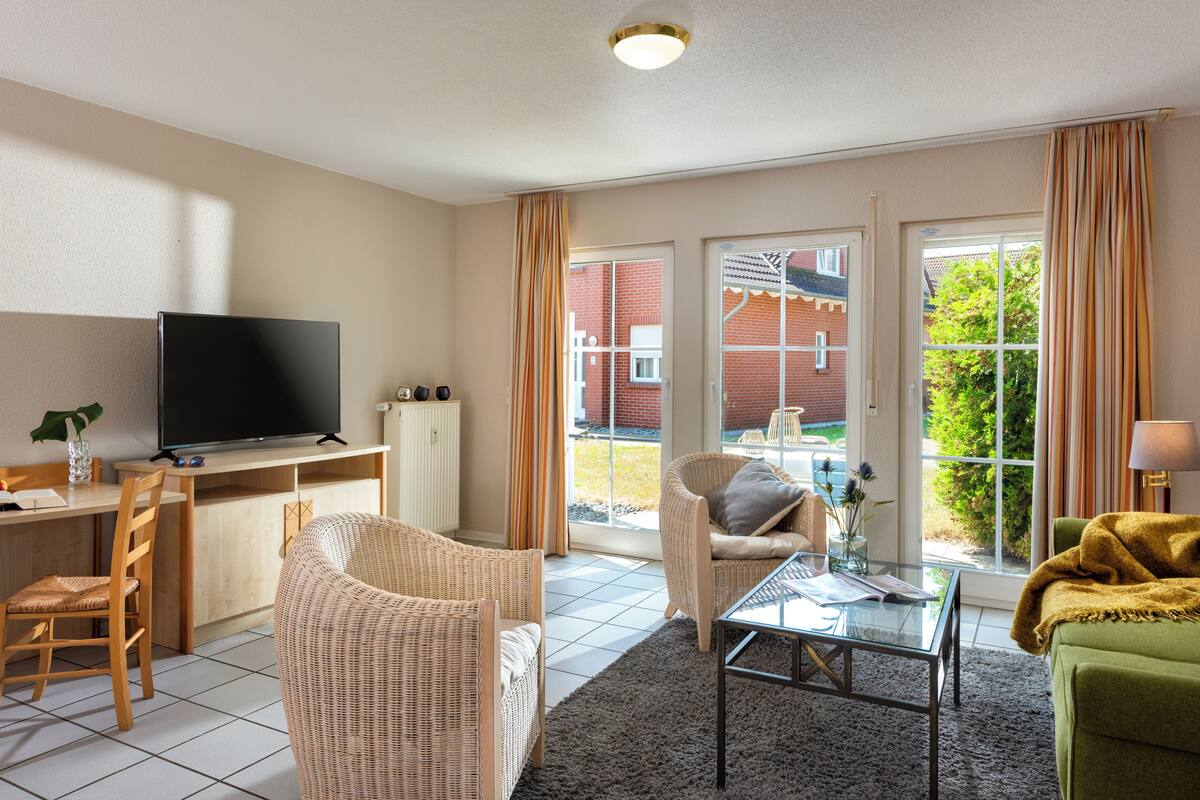 A modern living room with a flat-screen TV, wicker furniture, a glass coffee table, and a view of a brick building through large windows.