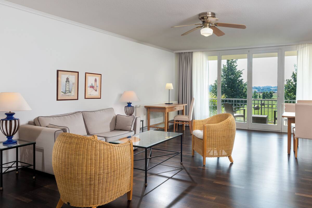 A spacious living room with a sofa, two wicker chairs, a glass coffee table, and a dining area with a table and chairs. There is a ceiling fan, large windows with curtains, and framed pictures on the wall.