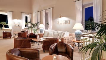 A spacious lounge area with leather sofas, armchairs, and a coffee table. There are potted plants and a lamp on a side table.