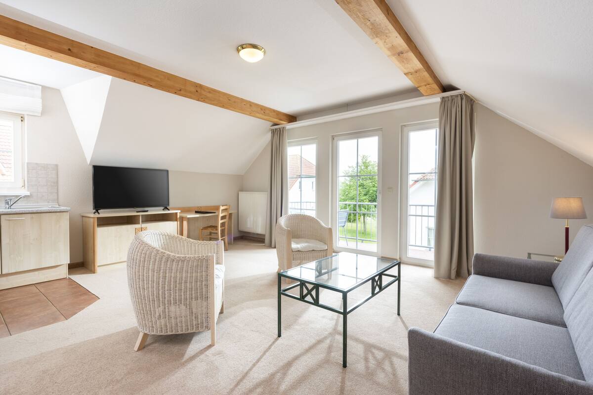 A spacious living room with a high ceiling, a flat-screen TV, a glass coffee table, a grey sofa, wicker chairs, and large windows offering a view of the outdoors.