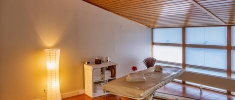 Una habitación con techo de madera, una camilla de masaje, una lámpara de pie y una ventana con persianas.