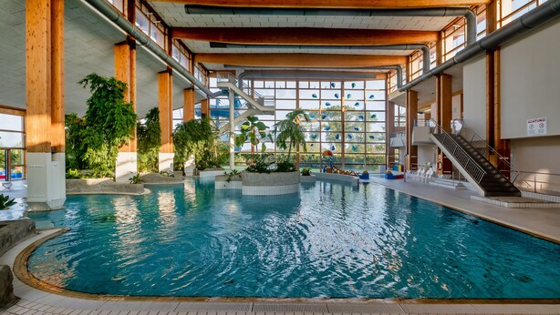 An indoor swimming pool with a central island, surrounded by plants and a staircase leading to upper levels. - Sunday Resort Rügen (Sagard)