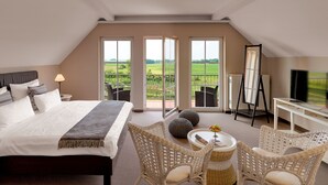 A hotel room with a large bed, a desk with a TV, wicker chairs, and a balcony with a view of the countryside. - Sunday Resort Rügen (Sagard)
