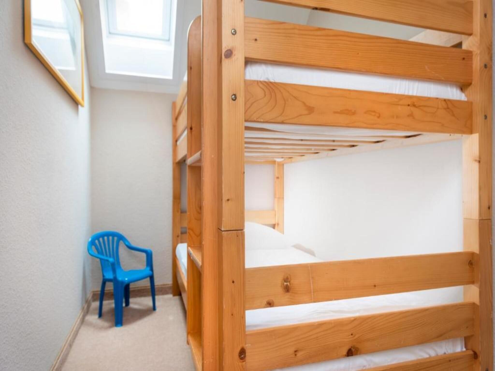 Un lit superposé avec une chaise bleue dans une pièce aux murs blancs et dotée d’une fenêtre.