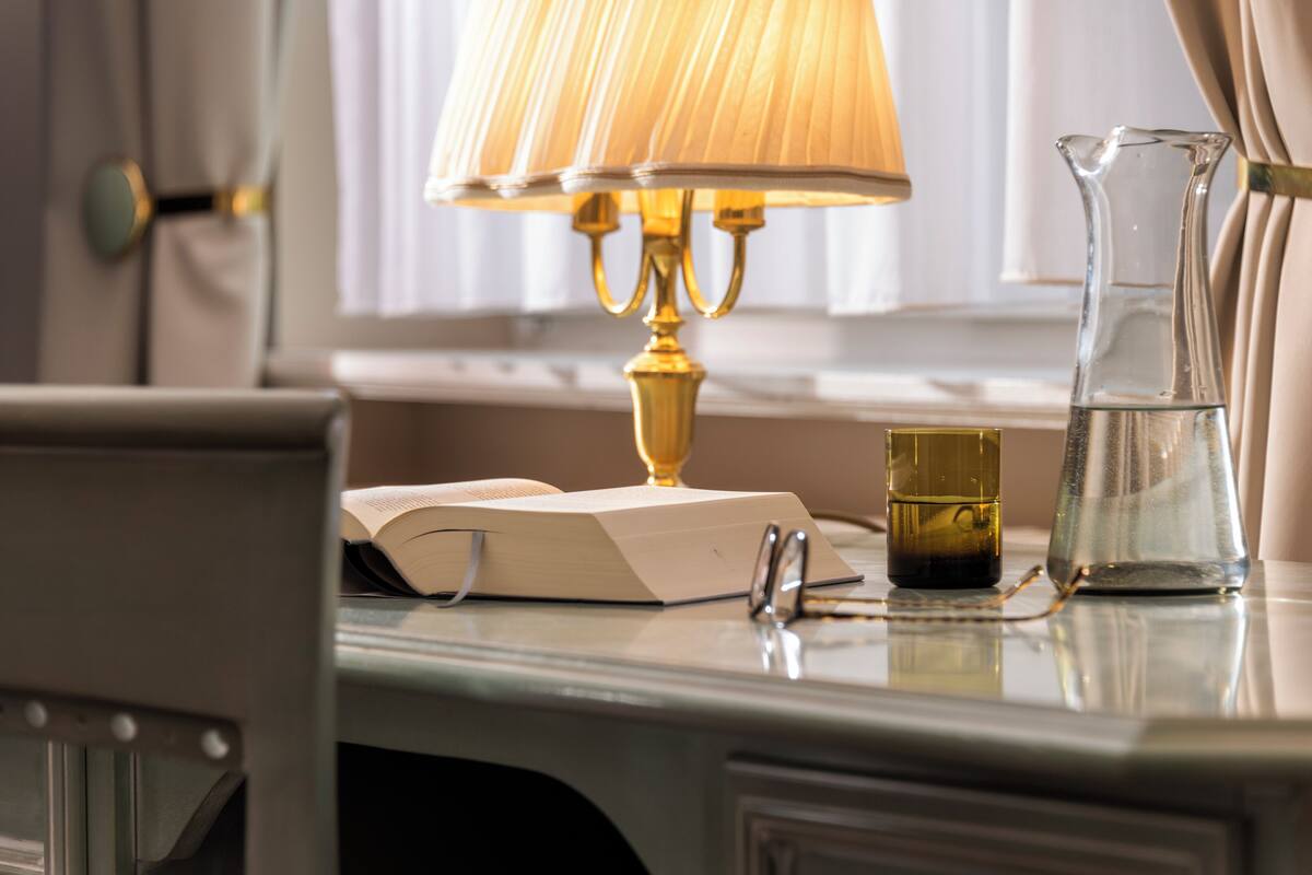 A desk with a lamp, a book, a glass pitcher, and a pair of glasses.
