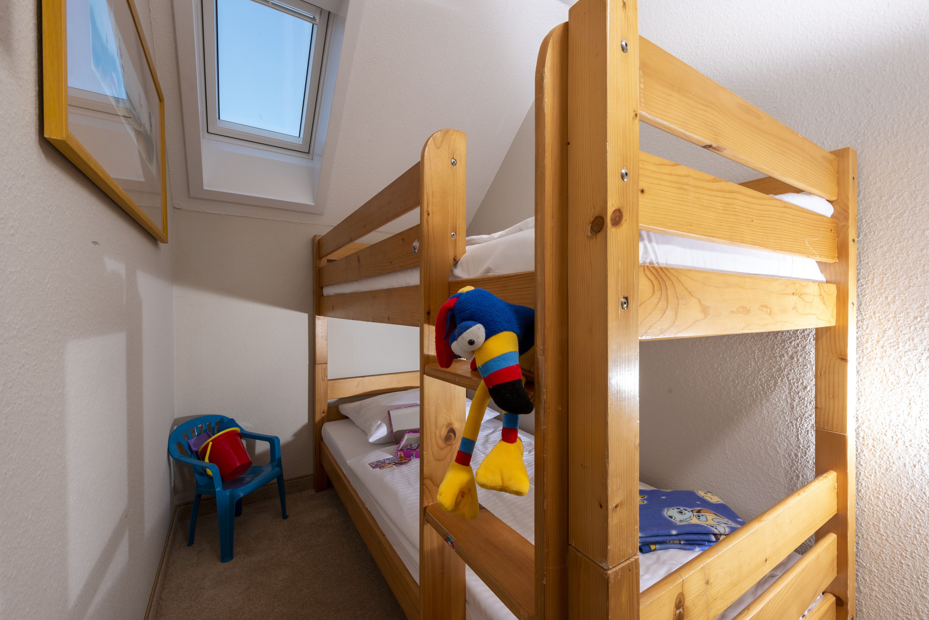 A bunk bed with a plush toy hanging from the top rail, a small blue chair, and a window above the bed.