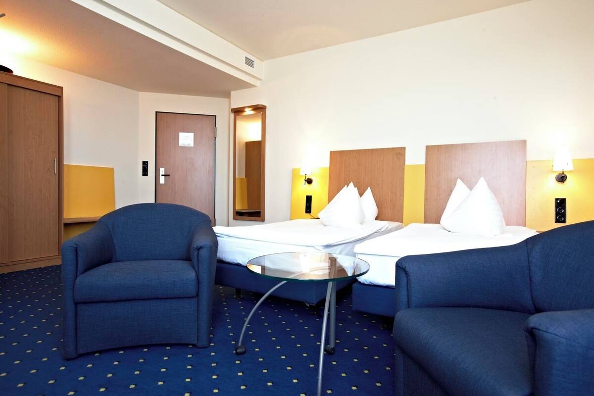 A hotel room with two beds, a small table, two armchairs, a wardrobe, and a mirror.