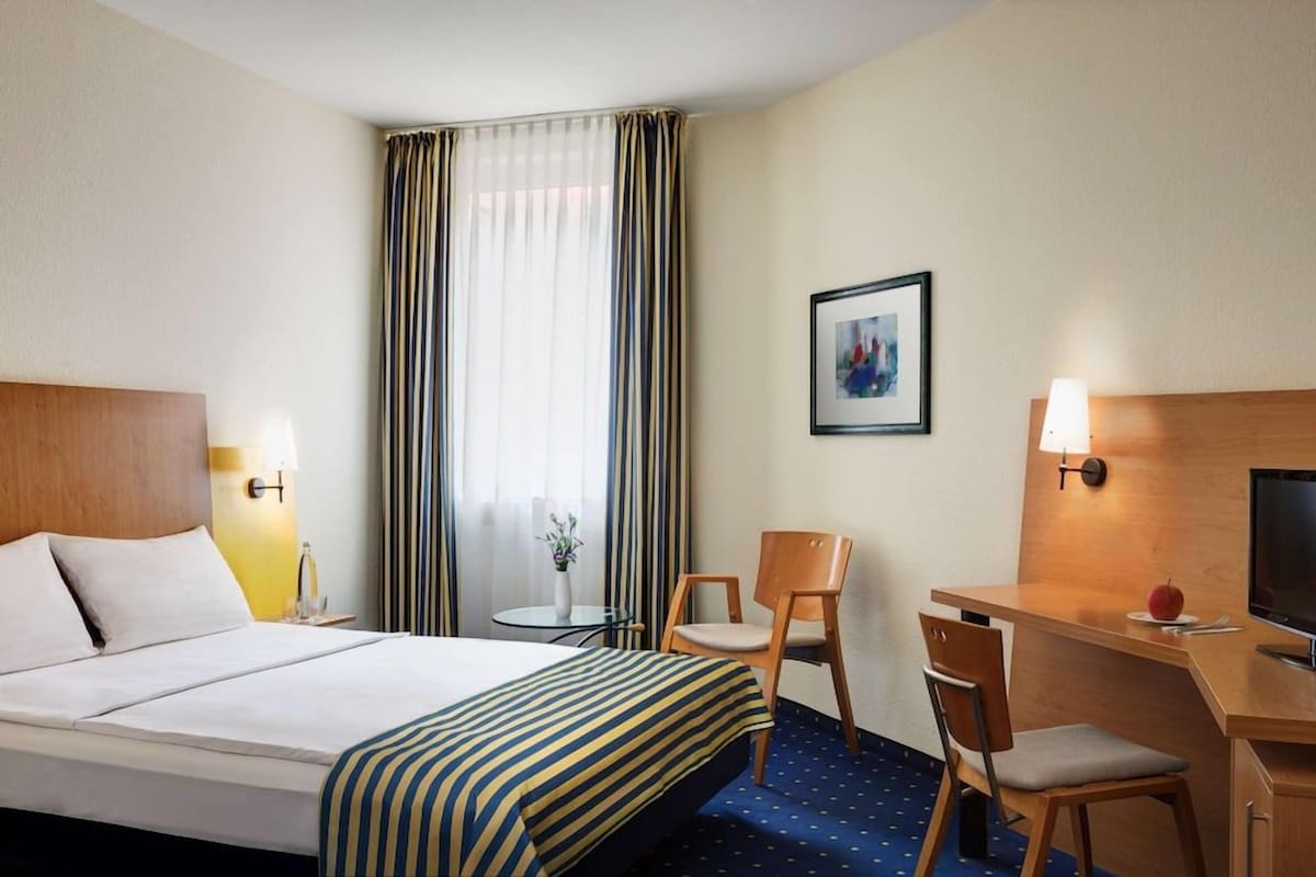 Habitación de hotel con cama, escritorio, silla, televisor, lámpara, un cuadro en la pared y una ventana con cortinas.