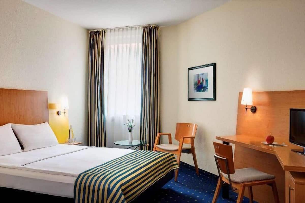 A hotel room with a bed, a desk, a chair, a television, a lamp, and a window with curtains.
