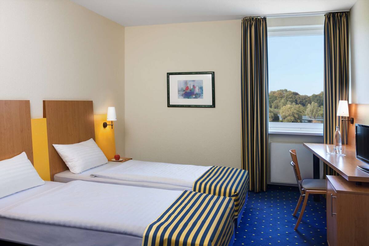 A hotel room with two beds, a desk, a chair, a TV, and a window with curtains.