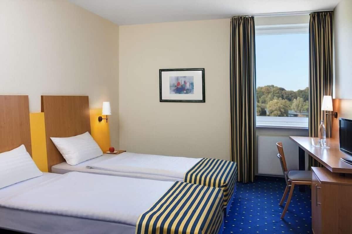 A hotel room with two beds, a desk, a chair, a TV, and a window with curtains.