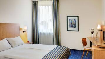 Een hotelkamer met een bed, een bureau, een stoel, een raam met gordijnen, en een ingelijst schilderij aan de muur.