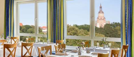 Un comedor con mesas dispuestas para cuatro, ventanas amplias con cortinas y vista a un edificio lejano.