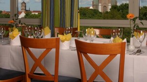 A dining area with a table set for two, featuring wooden chairs with blue cushions and a view of a church through large windows. - IntercityHotel Stralsund (Stralsund)