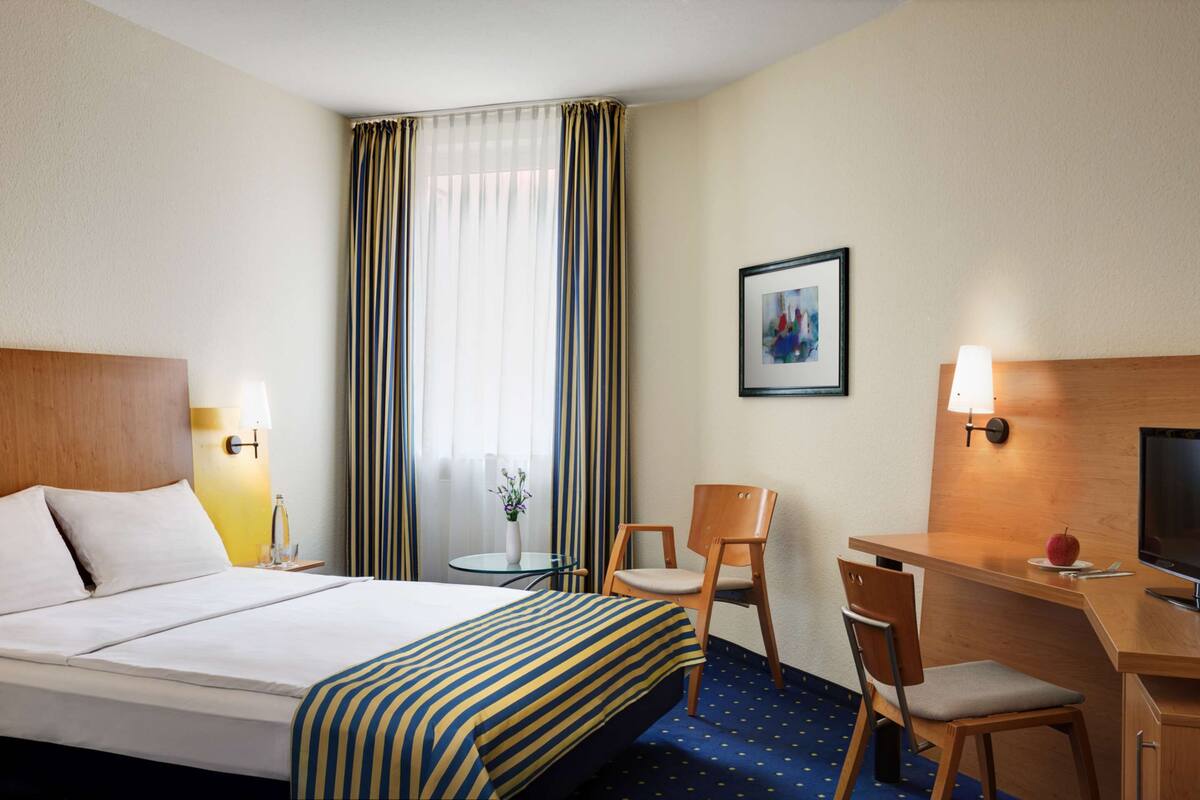 A hotel room with a bed, a desk with a chair, a television, a small table with a vase, and a window with curtains.